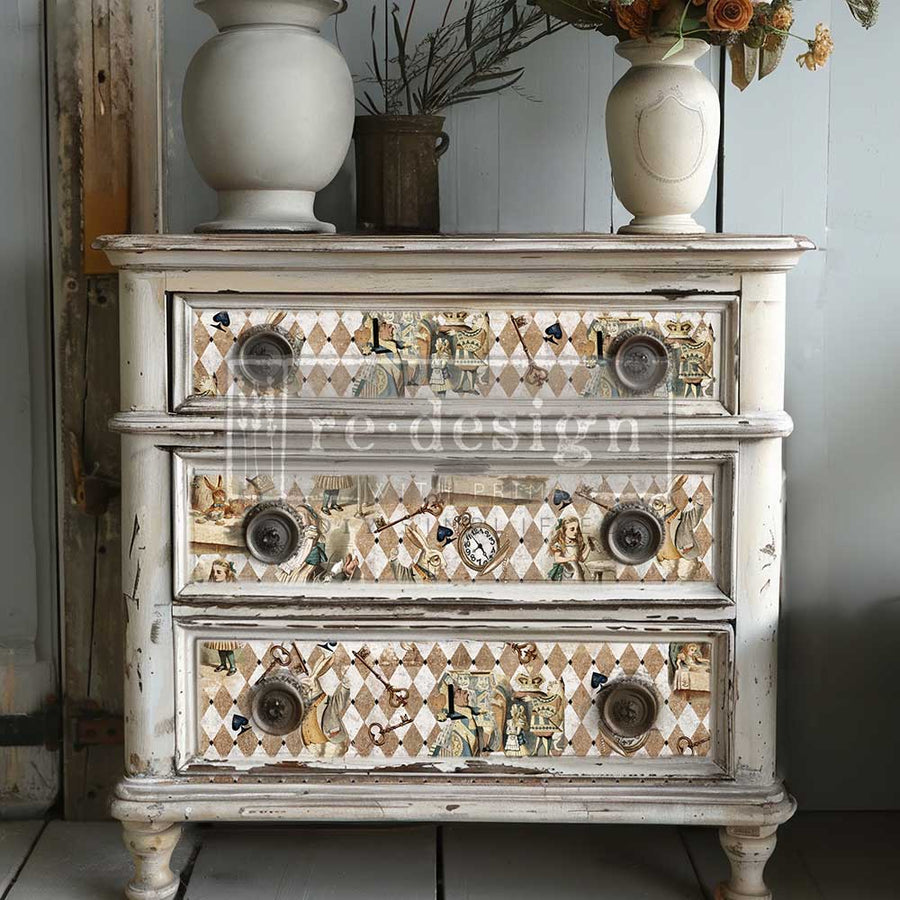 Distressed white dresser with all three drawers decorated in Redesign with Prima’s Mad Hatter’s Muse decoupage paper in a brown-and-beige Wonderland motif.